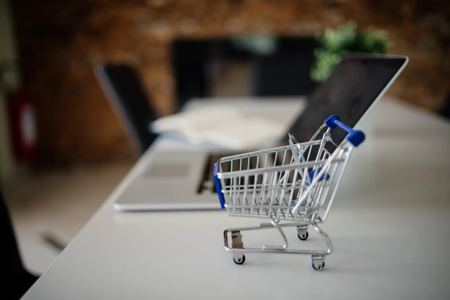 Miniature metal shopping cart on a white desk in front of a laptop, illustrating online shopping
